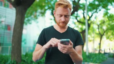 Parktaki bankta otururken akıllı telefondan mesaj atan dalgın kızıl saçlı adam.