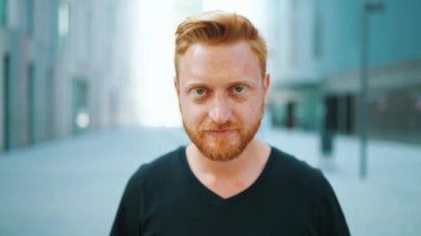 Mutlu kızıl saçlı adam şehirde dikilirken kameraya bakıyor.