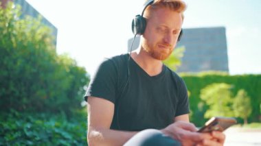 Pozitif kızıl saçlı adam kulaklıkla müzik dinliyor ve dışarıda bankta otururken telefonla yazıyor.