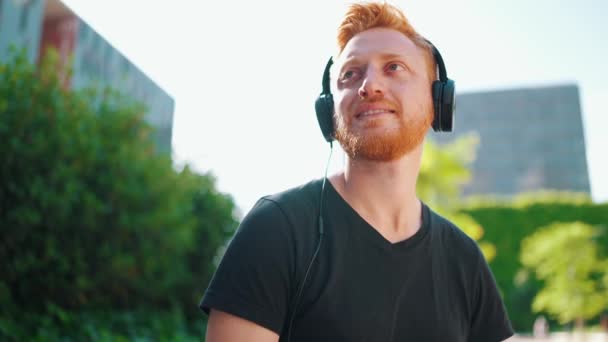 Χαρούμενος Κοκκινομάλλης Άνδρας Ακούει Μουσική Ακουστικά Ενώ Κάθεται Στον Πάγκο — Αρχείο Βίντεο