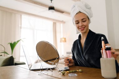 Beyaz kadın evde aynaya bakarken havluyu sarıp makyaj yapıyor.