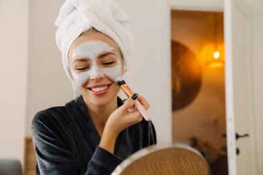 Beyaz kadın evde aynaya bakarken yüz maskesi takıyor.