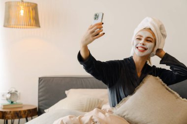 Evde yatarken cep telefonu kullanan maskeli genç bir kadın.