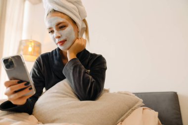 Evde yatarken cep telefonu kullanan maskeli genç bir kadın.