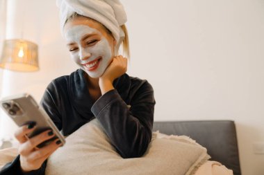 Evde yatarken cep telefonu kullanan maskeli genç bir kadın.