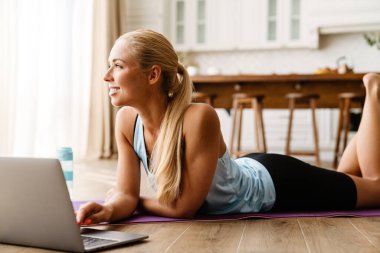 Evde yoga yaparken dizüstü bilgisayar kullanan sarışın genç bir kadın.