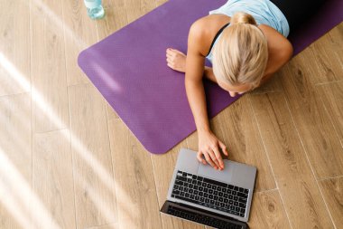 Evde yoga yaparken dizüstü bilgisayar kullanan sarışın genç bir kadın.