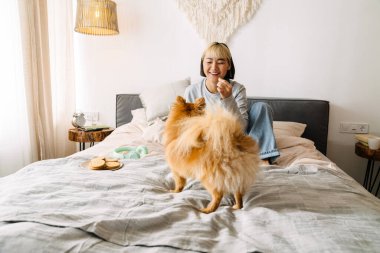 Asyalı bir kız evde oturmuş köpeğini beslerken gülüyor.