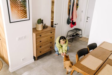 Evde çömelirken gülümseyen ve köpeğini besleyen Asyalı kız.