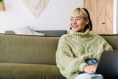 Asyalı kız evde kanepede dinlenirken bilgisayarla gülüp çalışıyor.