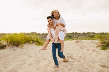 Yaz plajında sırtına binerken gülümseyen çok ırklı genç bir çift.