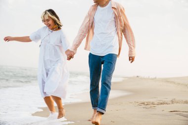 Yaz plajında yürürken gülümseyen ve el ele tutuşan çok ırklı bir çift.