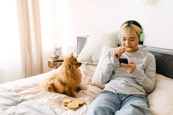 Asian girl eating cookie and using cellphone while resting with her dog on bed at home