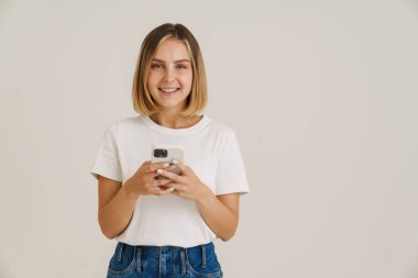 Beyaz arka planda izole edilmiş cep telefonu kullanırken gülümseyen genç sarışın kadın.