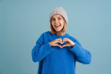 Mavi arka planda izole edilmiş kalp hareketi gösteren örgü şapkalı genç sarışın kadın.