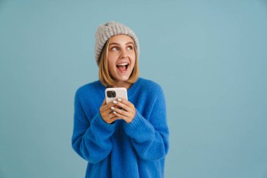 Mavi arka planda, örgü şapka takan, çığlık atan ve cep telefonu kullanan heyecanlı genç bir kadın.