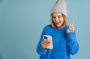 Örgü şapka takan genç sarışın kadın mavi arka planda izole edilmiş ve cep telefonu kullanıyor.