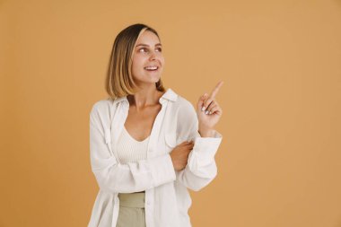Genç sarışın kadın gülümserken bej arka planda izole edilmiş bir şekilde parmağıyla işaret ediyor.