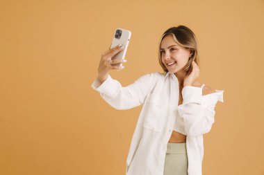 Bej arka planda izole edilmiş cep telefonuyla selfie çekerken gülümseyen genç sarışın kadın.