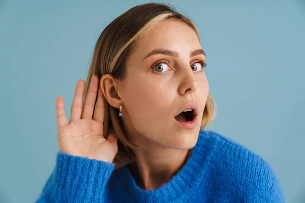 Young shocked woman wearing sweater gesturing and looking at camera isolated over blue background