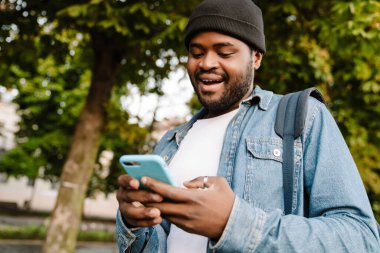 Siyah genç adam yaz parkında yürürken cep telefonu kullanıyor.