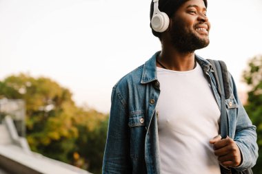 Şapkalı siyahi adam sonbahar parkında yürürken kulaklıkla müzik dinliyor.