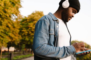 Sonbahar parkında yürürken kulaklık takan siyah bir adam cep telefonu kullanıyor.