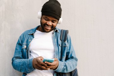 Kulaklıklı, gülümseyen ve dışarıda cep telefonu kullanan siyahi bir genç.