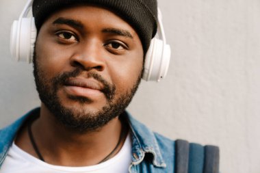 Şapkalı genç adam kulaklıkla dışarıda müzik dinliyor.