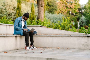 Parkta bankta otururken dizüstü bilgisayarla çalışan siyahi bir genç.
