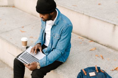 Kulaklıklı siyah bir adam dışarıda bankta otururken dizüstü bilgisayarla çalışıyor.