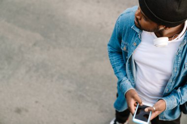 Kulaklıklı siyahi bir adam dışarıda beklerken cep telefonu kullanıyor.