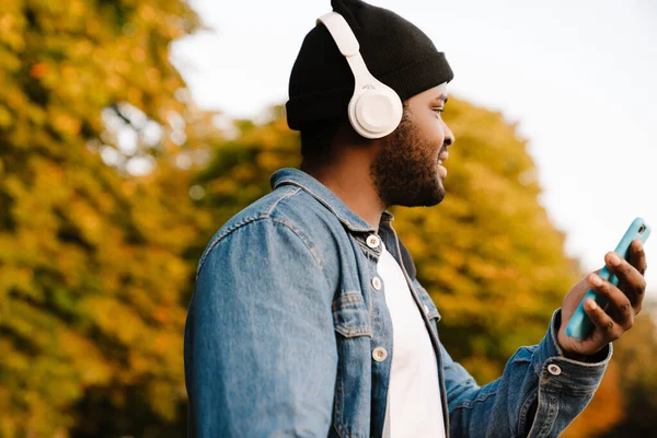 秋の公園を歩いている間 携帯電話を使ってヘッドフォンで黒人男性 — ストック写真