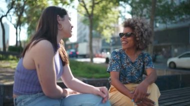 İki gülümseyen etnik kadın bankta otururken konuşuyor.
