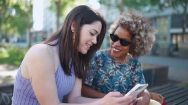 İki neşeli çoklu etnik kadın dışarıda bankta oturmuş telefonla konuşuyor ve bakıyor.