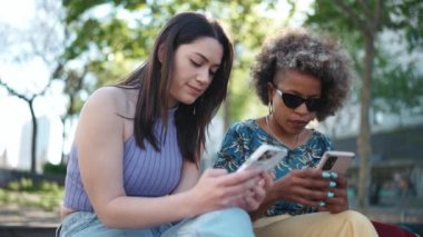 İki yakışıklı çoklu etnik kadın dışarıda bankta oturmuş telefonla konuşuyor ve bakıyor.