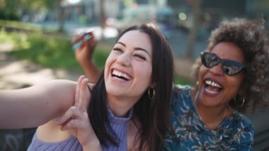İki tane gülümseyen etnik kadın dışarıda bankta otururken telefonla selfie çekiyorlar.