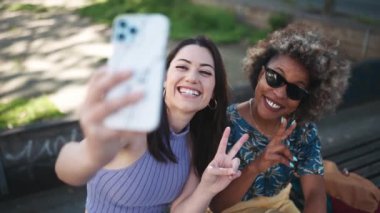 İki neşeli çoklu etnik kadın dışarıda bankta otururken telefonla selfie çekiyorlar.