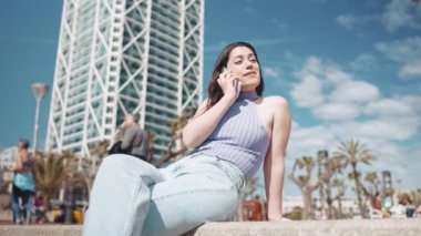 Neşeli esmer kadın dışarıda bankta otururken telefonla konuşuyor.