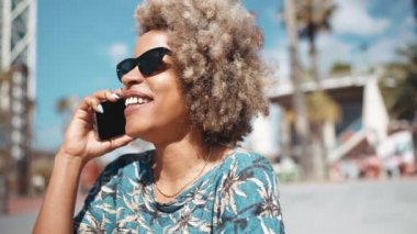 Gülümseyen Afrikalı gri saçlı, güneş gözlüklü, açık hava bankında cep telefonuyla konuşan kadın.