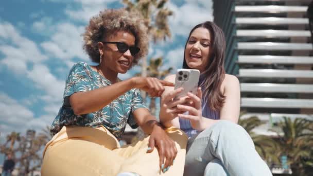 Uśmiechnięte Słoneczne Wieloetniczne Kobiety Rozmawiające Patrzące Telefon Siedząc Zewnątrz — Wideo stockowe