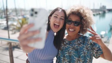 Çok ırklı iki kadın, yatların yanındaki sette telefonda selfie çekiyorlar.