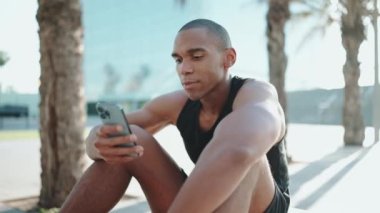 Siyah tişört giyen kel Afrikalı sporcu sabahları dışarıda telefonla yazı yazıyor.