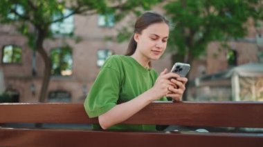 Yeşil tişört giyen, dışarıda bankta telefonla mesajlaşan mutlu sarışın kadın.