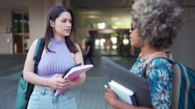İki gülümseyen çoklu etnik öğrenci ellerinde kitaplarla konuşan kadınlar.