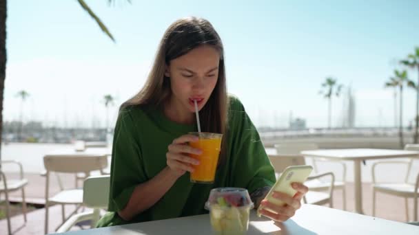 真剣なブロンド女性身に着けています緑のTシャツ見ます電話と飲みオレンジジュースでカフェ屋外 — ストック動画