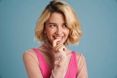 Sarı saçlı genç beyaz kadın gülüyor ve mavi duvarda parmağını ısırıyor.