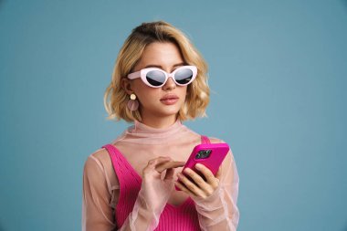 Mavi duvarda izole edilmiş cep telefonu kullanan güneş gözlüklü genç beyaz kadın.