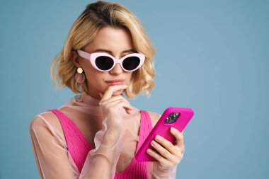 Mavi duvarda izole edilmiş cep telefonu kullanan güneş gözlüklü, kafası karışmış genç bir kadın.