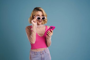 Mavi duvardan izole edilmiş cep telefonu kullanan genç beyaz kadın şaşkınlığını dile getiriyor.